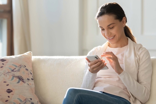 Lächelnde Frau sitzt auch Couch und tippt etwas auf ihrem Smartphone.